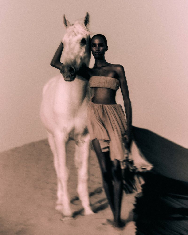An adult woman with dark skin stands next to a white horse on a sand dune in a beauty and fashion editorial shot with warm lighting.