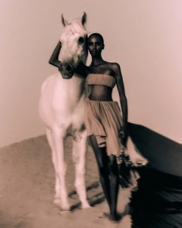 An adult woman with dark skin stands next to a white horse on a sand dune in a beauty and fashion editorial shot with warm lighting.