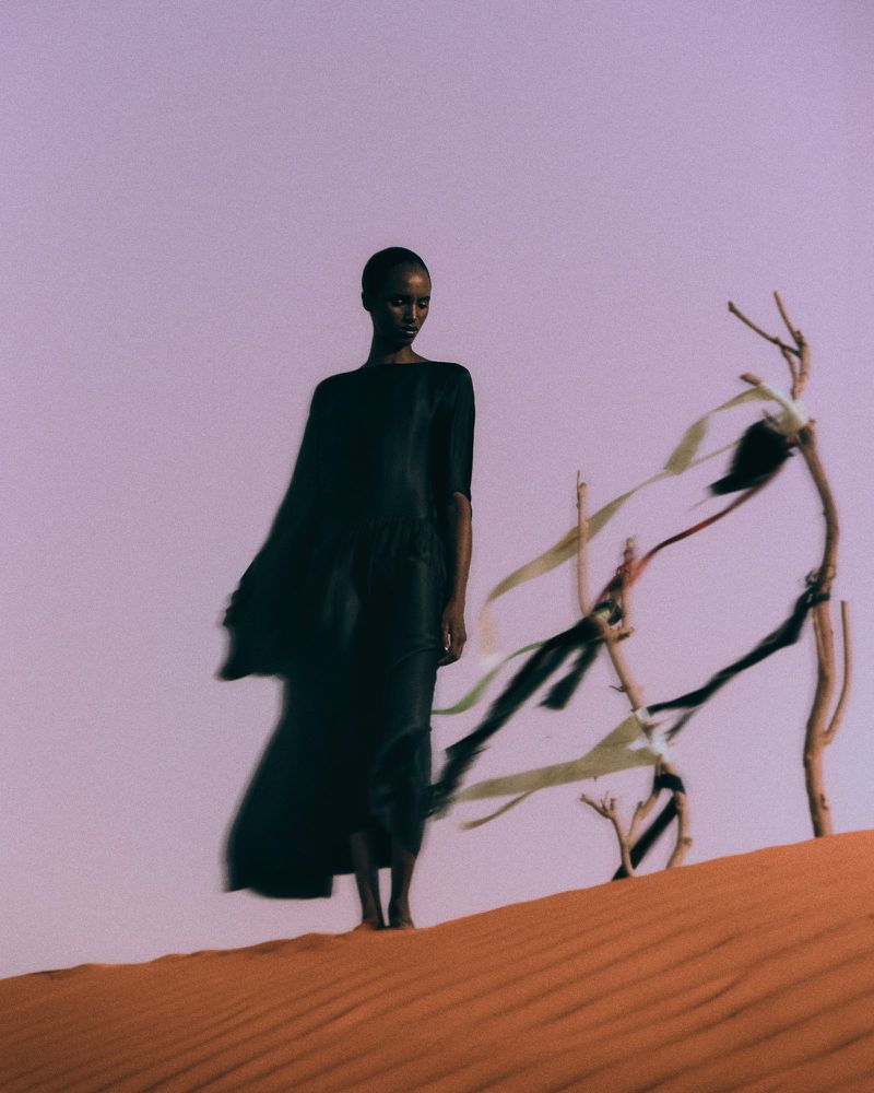 Woman in flowing black dress stands on sand dune next to abstract branches against a light purple sky in a fashion editorial photograph.