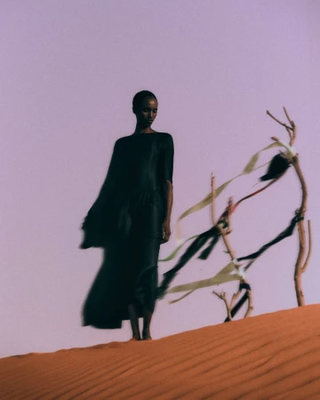 Woman in flowing black dress stands on sand dune next to abstract branches against a light purple sky in a fashion editorial photograph.