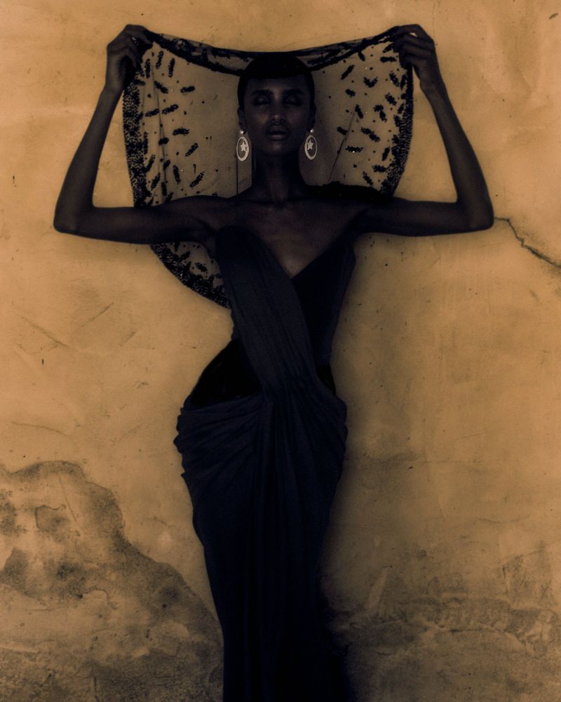 A woman in a draped dress and beaded head covering poses in front of a textured golden-beige wall for a fashion or beauty editorial.