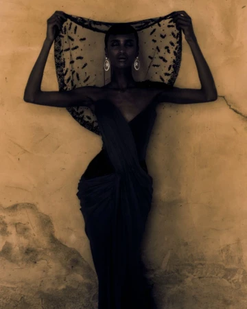 A woman in a draped dress and beaded head covering poses in front of a textured golden-beige wall for a fashion or beauty editorial.