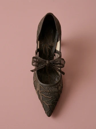 Top-down view of a dark brown lace high-heeled shoe featuring an embellished bow on a solid pinkish-brown studio background.