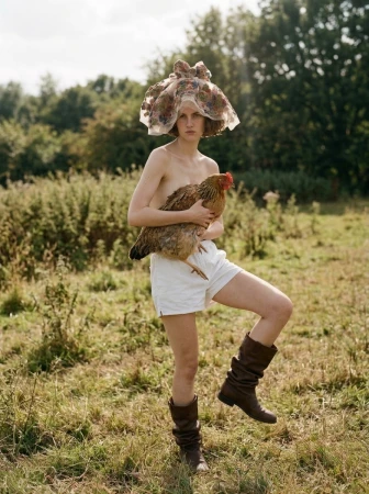 Model wearing a sheer floral headpiece and brown boots standing in a grassy field holding a live chicken.