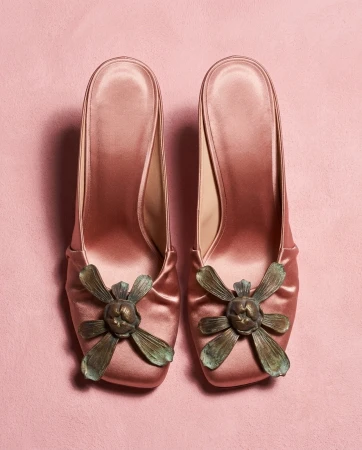 Top-down view of pink satin high-heel mules with sculptural bronze flower ornaments on a pink textured background.