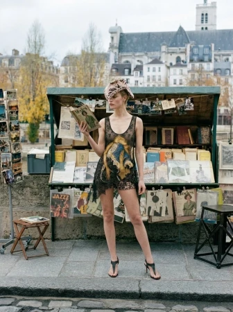 Model wearing a black lace dress and patterned hat reading a book in front of a riverside book stall in Paris