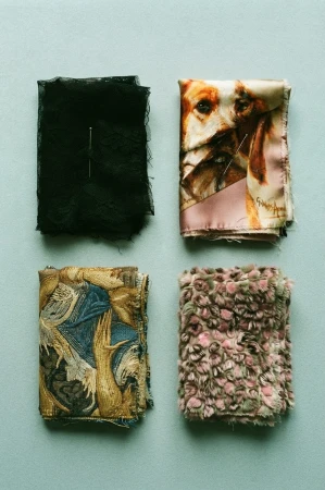 Four folded fabric swatches including black lace, dog print silk, embroidered tapestry, and textured pink faux fur on sage background