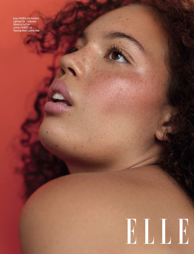 Close-up studio shot of a fair-skinned young adult female model with curly hair, freckles, and pink blush, looking up against an orange background.