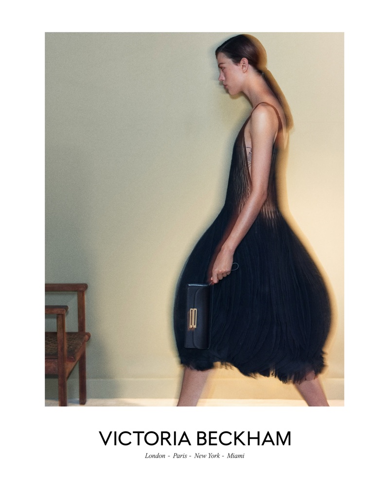 Model in a black pleated dress holding a dark clutch walking past a wooden chair against a neutral wall for Victoria Beckham.