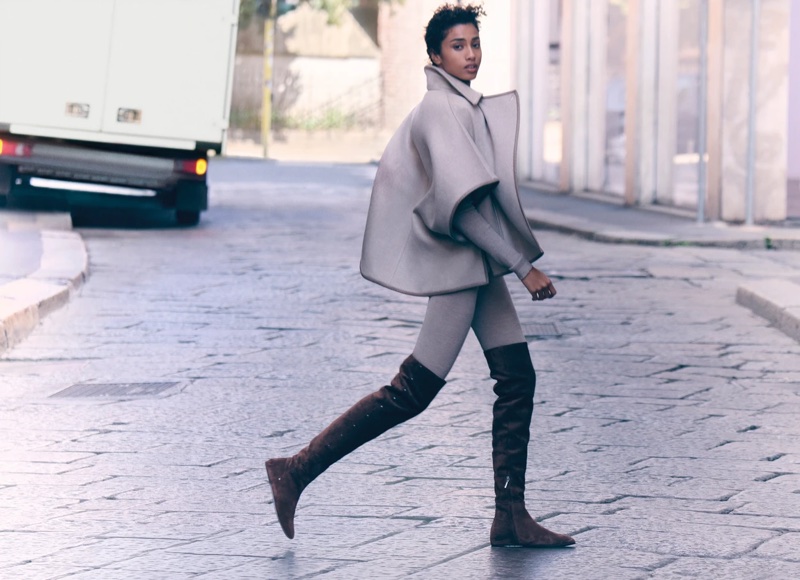 Model Imaan Hammam walking on a cobblestone street wearing a grey coat and tall brown boots for Max Mara
