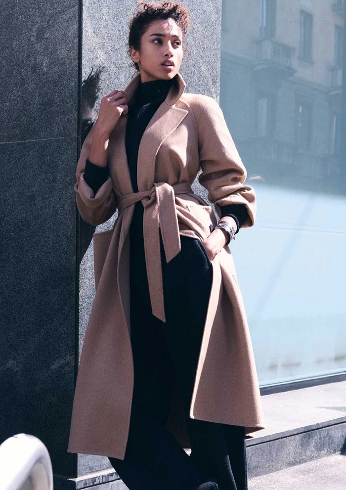 Model Imaan Hammam wearing a belted camel coat and black turtleneck leaning against a stone building exterior.