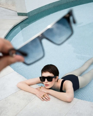 Anouk Weishaupt wearing black sunglasses in a swimming pool while a pair of sunglasses is held out of focus in the foreground.