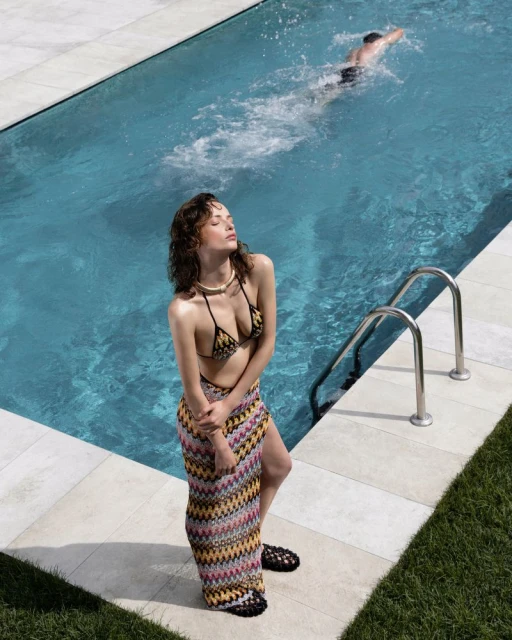 A woman in swimwear and a skirt poses by a pool on a sunny day, with a swimmer visible in the background, creating a summer fashion image.