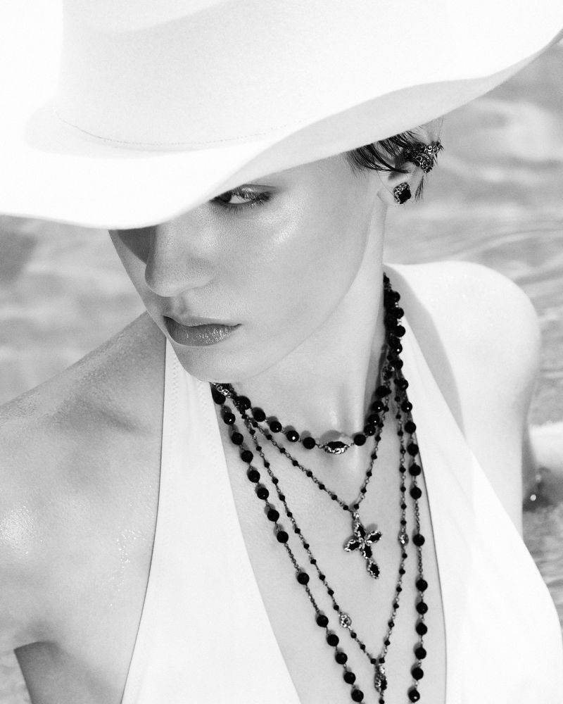 Monochrome portrait of a woman wearing a wide-brimmed hat and layered necklaces, gazing at the camera in a soft outdoor light.