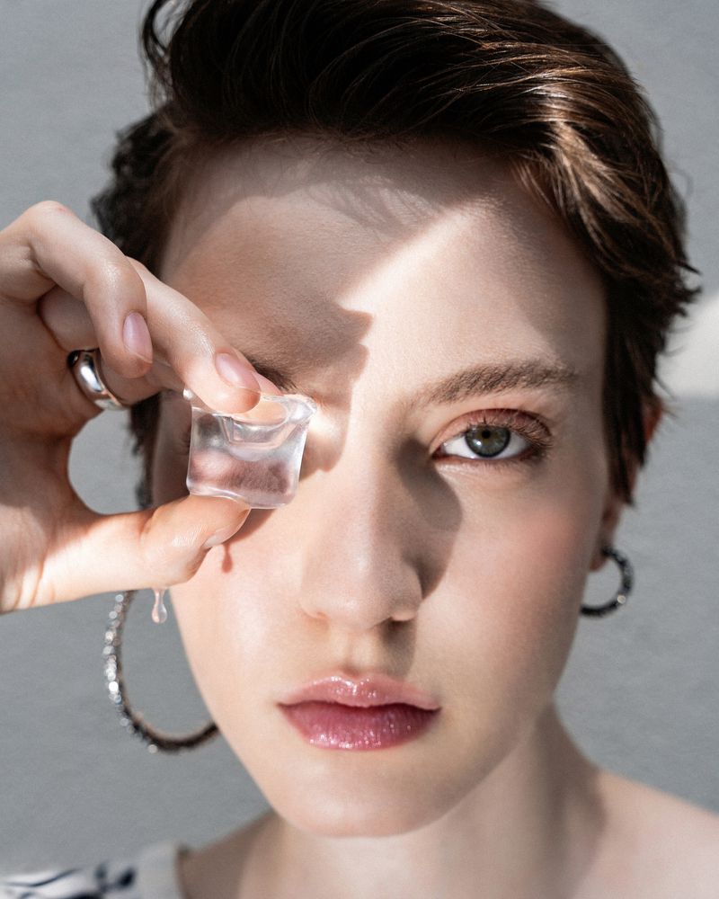 Close-up of a woman with short brown hair holding an ice cube to her face, wearing silver hoop earrings, in a beauty shot.