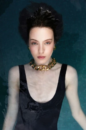 Woman with dark hair and fair skin in a black swimsuit wearing a gold chain in water against a dark teal background.