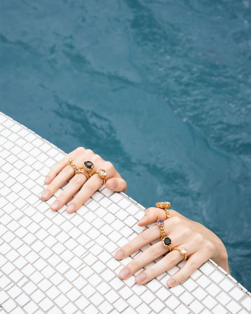 Hands adorned with gold rings, showcasing various gemstones, reaching over the edge of a mosaic-tiled pool with blue water.