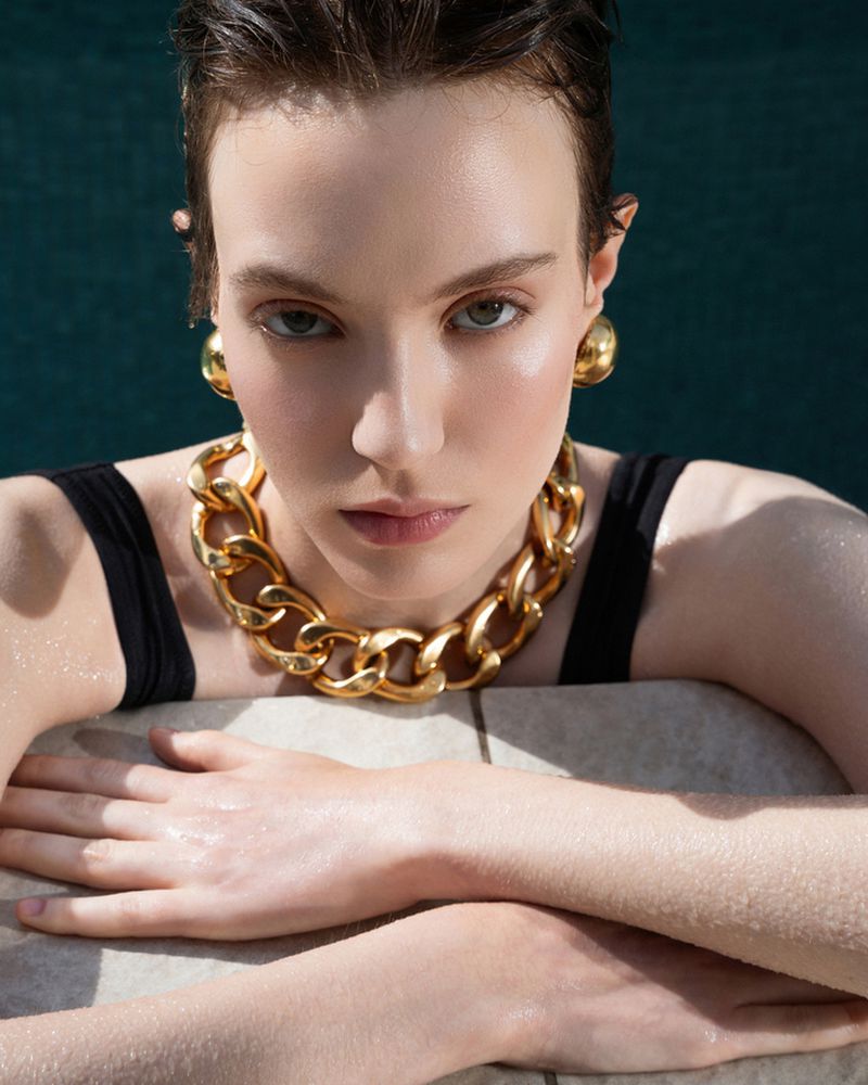 Close-up of a young adult female model with gold jewelry, wet hair, and black tank top, resting her arms, with a teal background.