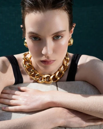 Close-up of a young adult female model with gold jewelry, wet hair, and black tank top, resting her arms, with a teal background.