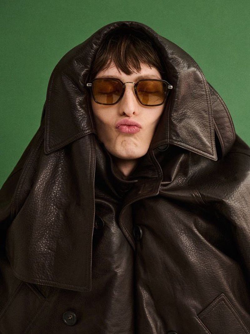 An adult man with sunglasses pouts at the camera, wearing an oversized brown leather jacket with a green backdrop for a studio shot.