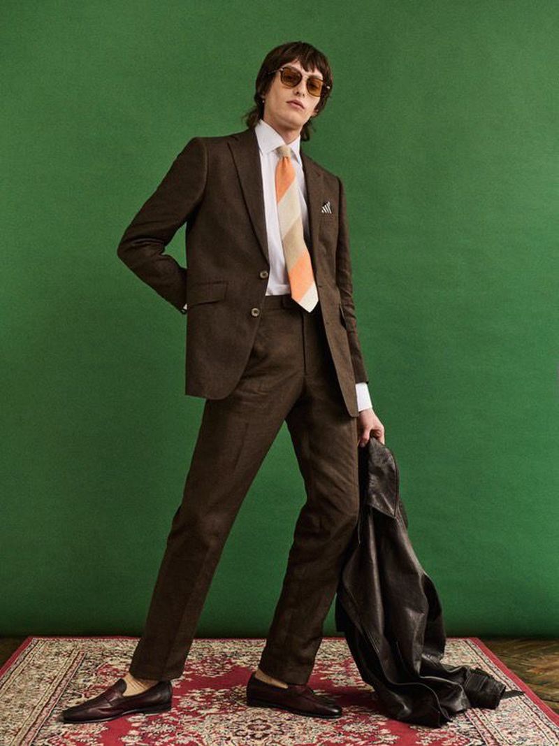 A full shot of a male model posing in a brown suit with striped tie and sunglasses on a red patterned carpet in front of a green backdrop.