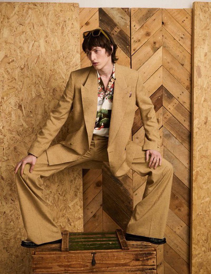 A male model in a tan suit poses atop a wooden box against a wood panel background, wearing sunglasses and a patterned shirt.