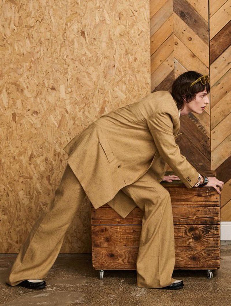 A slender adult male model poses crouching on a wooden box with a light-brown suit in a studio with wooden backdrops, fashion shot.