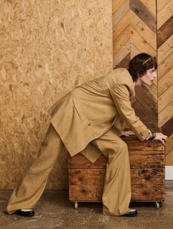 A slender adult male model poses crouching on a wooden box with a light-brown suit in a studio with wooden backdrops, fashion shot.
