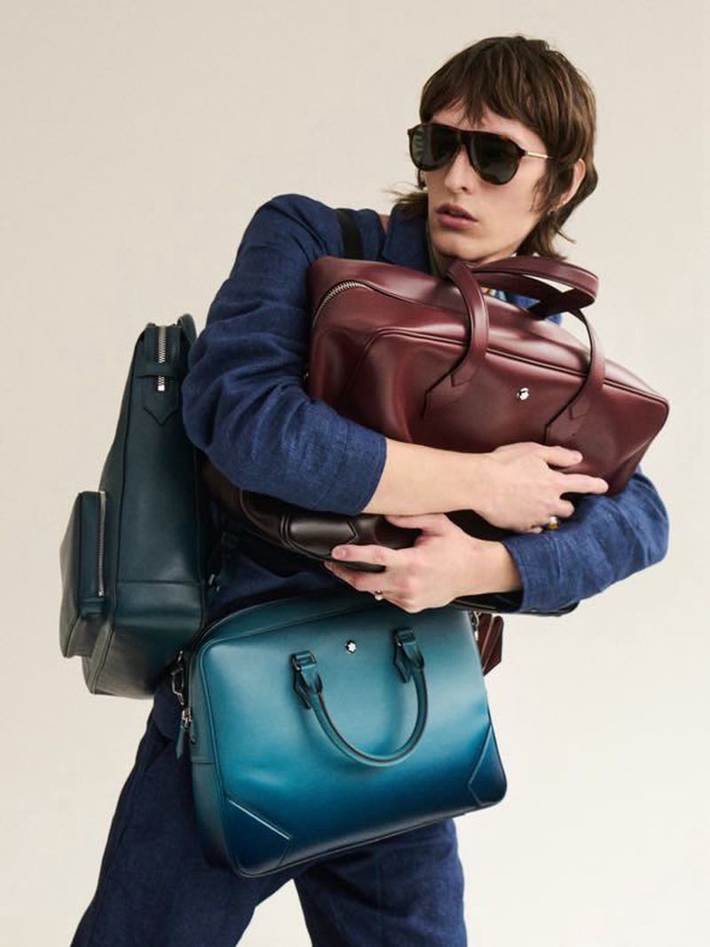 A person wearing sunglasses carries burgundy and teal Montblanc leather bags in a studio shot on a light-beige background.