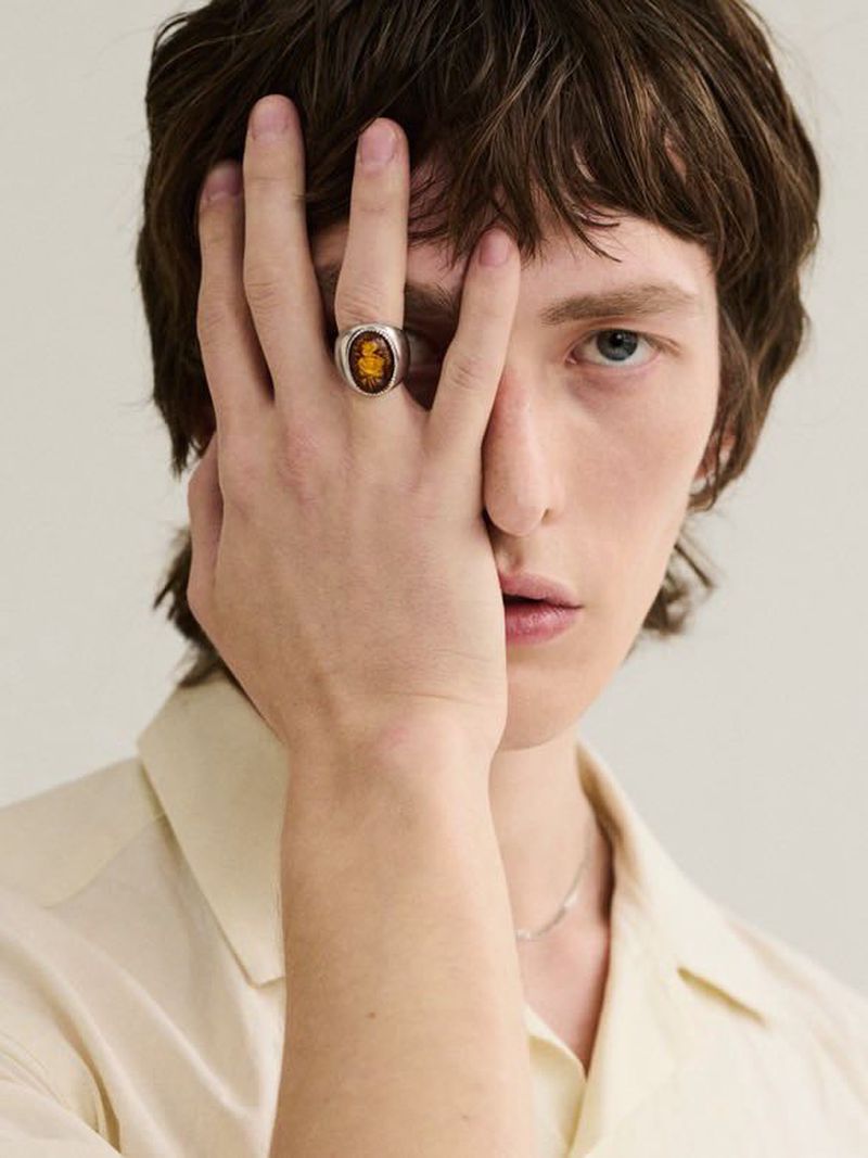 Close-up of a fair-skinned male model with an amber ring on his hand obscuring part of his face. He's wearing a cream-colored collared shirt.