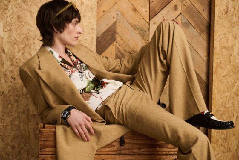 Man in retro fashion suit, floral shirt, sunglasses, and loafers lounging on wooden structure in front of matching wood wall.