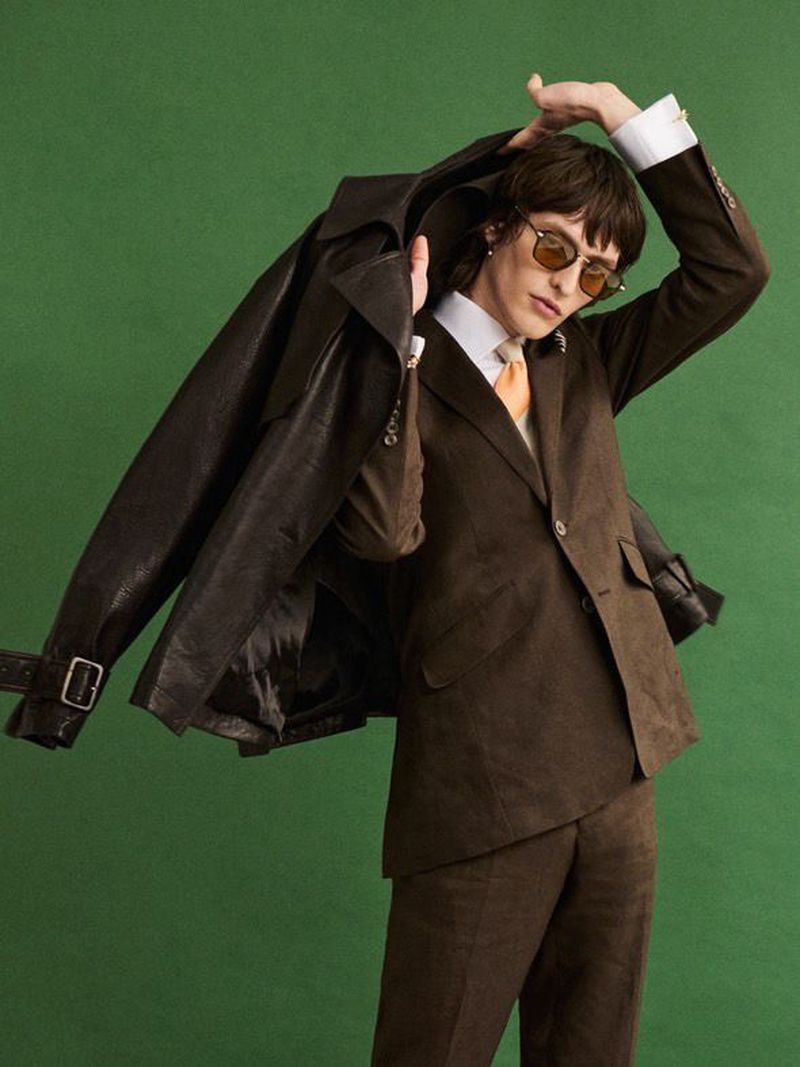 A person wearing sunglasses and a brown suit with a leather jacket draped over their arm poses against a solid green backdrop.