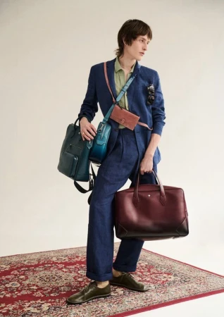 Man in a blue suit holding a burgundy briefcase, a backpack, and other bags in a studio on a patterned red rug.