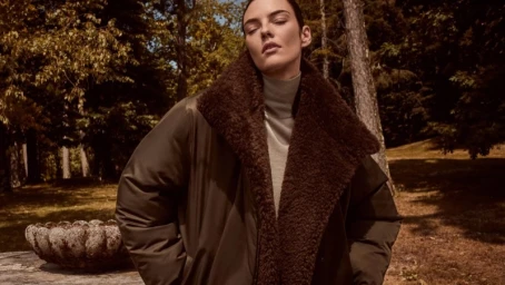 Model in a dark brown coat with a furry collar outdoors among trees for the Vince Fall 2026 campaign.