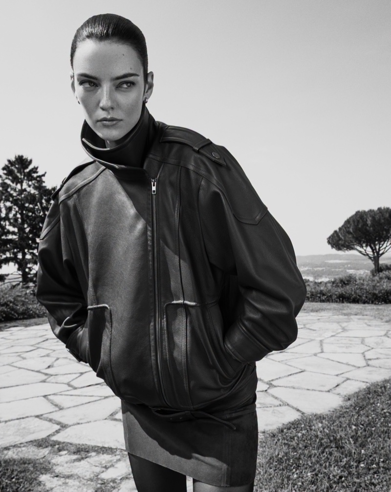 Model wearing a dark oversized leather jacket outdoors against a sunny terrace and landscape background