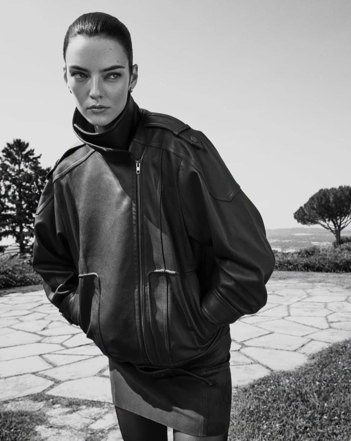 Model wearing a dark oversized leather jacket outdoors against a sunny terrace and landscape background