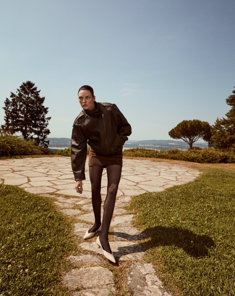 Model wearing a dark leather jacket and skirt on a stone path outdoors under a clear sky for Vince.