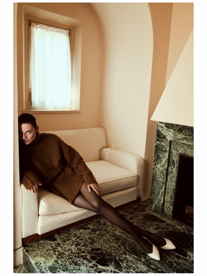Model wearing brown knit sweater and black tights reclining on a white sofa next to a marble fireplace
