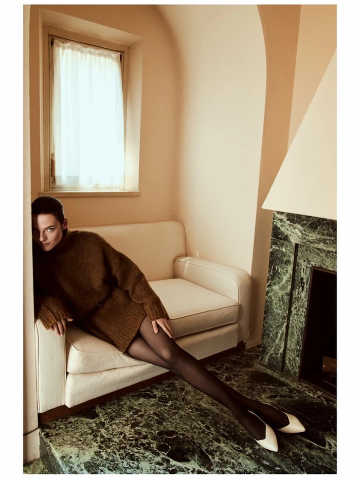 Model wearing brown knit sweater and black tights reclining on a white sofa next to a marble fireplace