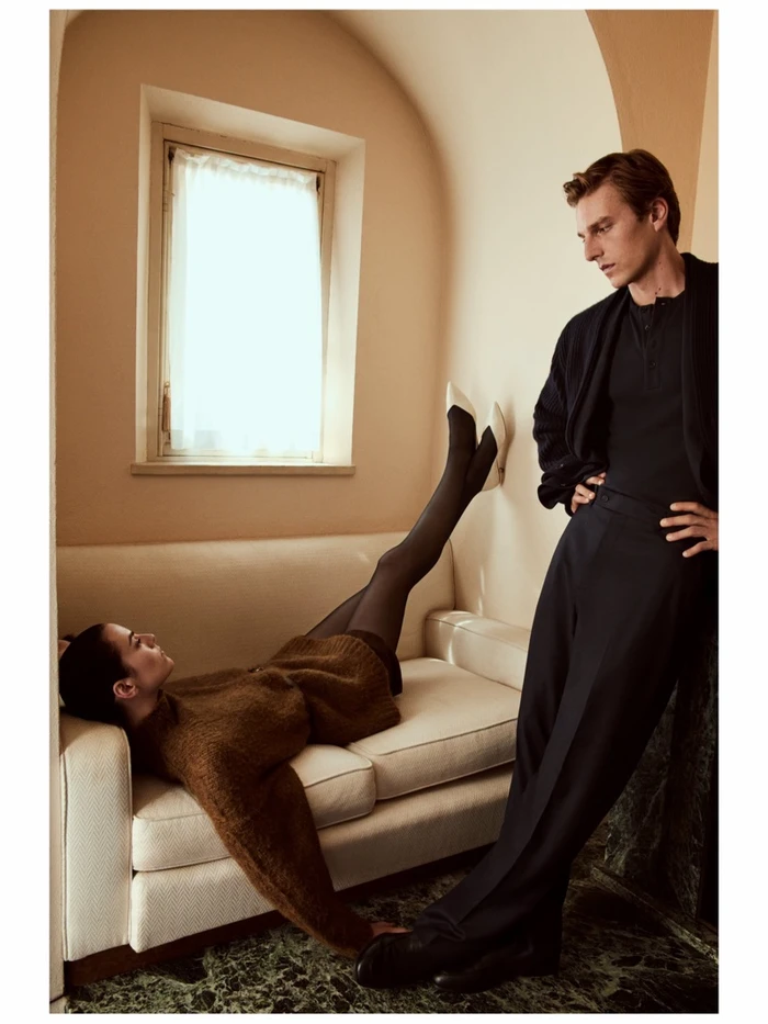 Woman lying on a cream sofa with legs up against a wall, while a man stands beside her in an interior room with a window.