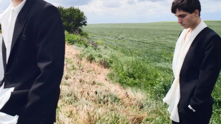 Two men in dark suits standing outdoors in a grassy rural landscape with an overcast sky.