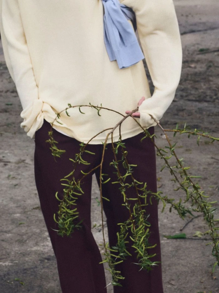 Close-up of a person wearing a cream sweater, light blue scarf, and dark trousers holding a small tree branch with green catkins.