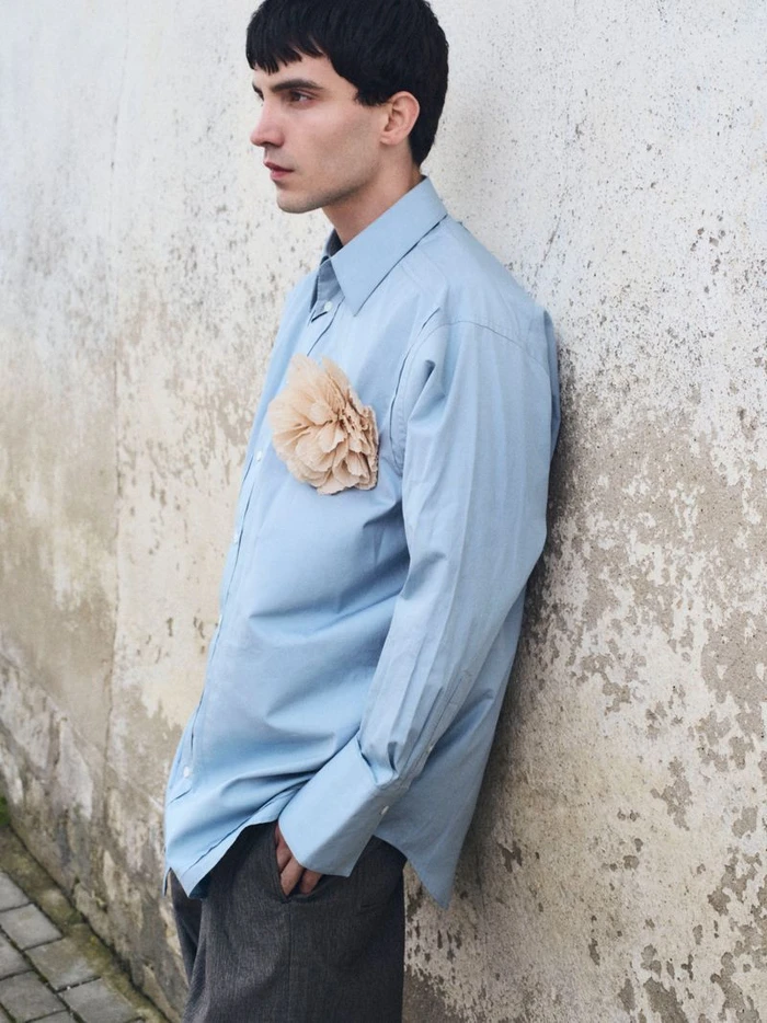 Man wearing light blue button-down shirt with a fabric flower pin leaning against a textured wall for Teeth Magazine