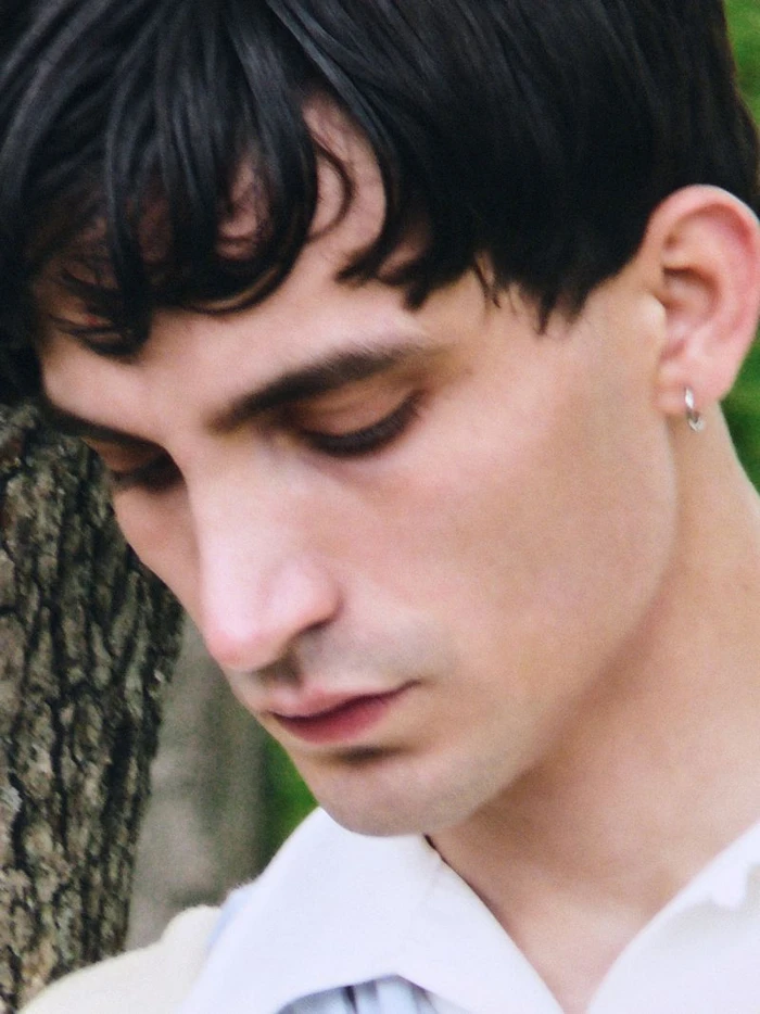 Close-up portrait of a young man with dark hair looking down, wearing a white shirt and a small hoop earring, outdoors near a tree.