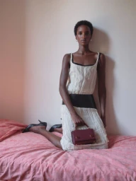 Model kneeling on a pink bed holding a small maroon leather handbag, wearing a white textured dress with a black waist belt.