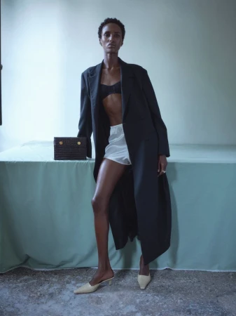 Model standing behind a table draped in sage green cloth, wearing a long black coat, white shorts, and holding a dark handbag.