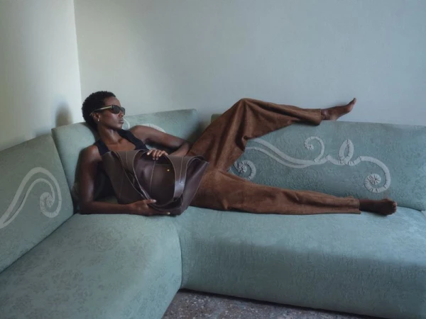 Model wearing dark sunglasses and brown suede trousers reclining on a light green patterned sofa holding a brown leather bag