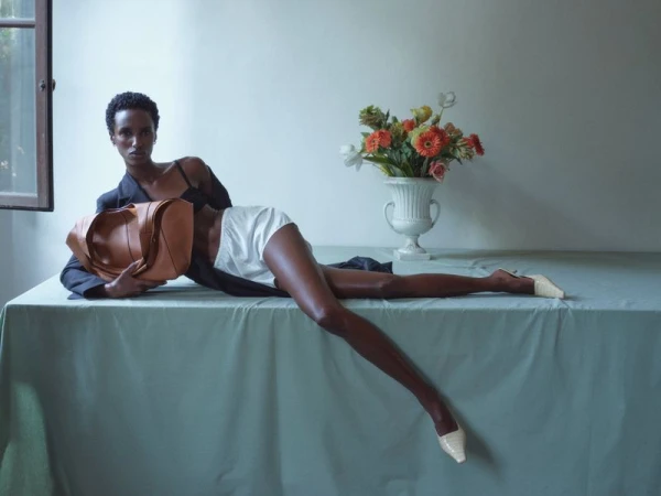 Model reclining on a cloth-covered table holding a tan leather bag beside a vase of flowers in an editorial portrait