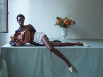 Model reclining on a cloth-covered table holding a tan leather bag beside a vase of flowers in an editorial portrait