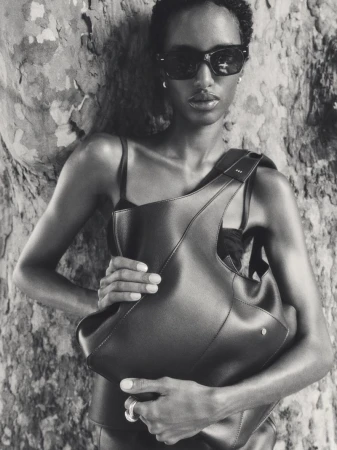 Black and white fashion portrait of a model wearing sunglasses and holding a black leather handbag against a textured tree trunk.